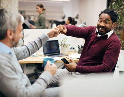 Two smiling coworkers fist bumping while sitting at their desks.